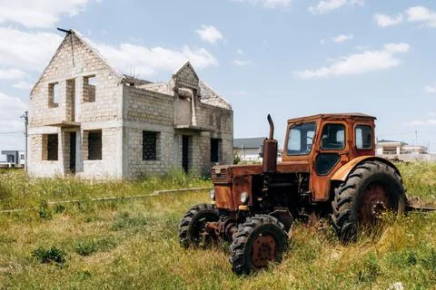 Rusty old tractor standing in overgrown field beside unfinished stone house Stock Photos