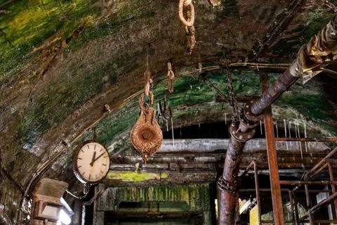 Rusty pulley, chains, pipes, clock, and lamp hang beneath damp brick ceiling  Stock Photos