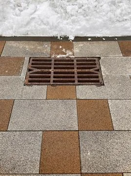 Rusty storm drain grate on tiled road and snowdrift that will melt in spring Stock Photos