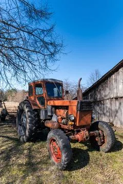 Rusty tractor is stationed in the yard alongside a weathered barn and trees. Stock Photos