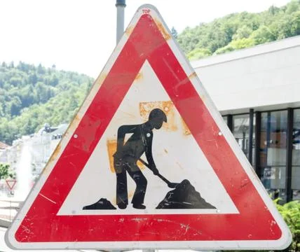 A rusty triangular sign warning of roadworks ahead with a modern city Stock Photos