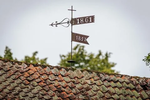A rusty weather vane with dates 1861 and 1857 stands atop a mossy tiled roof Foto stock