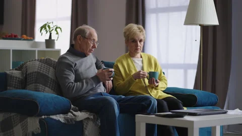 Sad aged spouses are sitting at home and drinking tea in living room, talking to Stock Footage 153610120