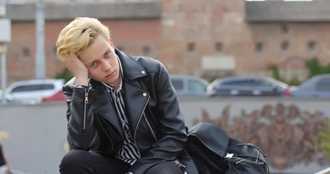Sad attractive blond guy sitting on a bench in the city and resting his head on Stock Footage 126576153