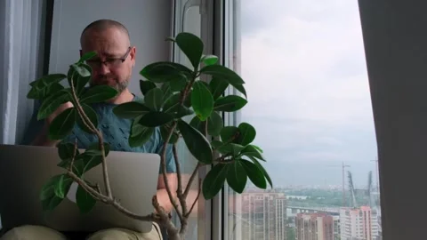 Sad bearded man in glasses using a laptop while sitting on the windowsill. Stock Footage 246890945