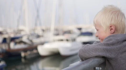 A sad blond boy stands on the pier near the yachts and looks at the sea Stock Footage 108463350
