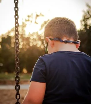 Sad boy with glasses looks at the floor sitting on a swing without playing Stock Photos