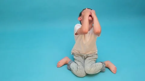 Sad boy sitting on floor covering his face with hand. Upset child expressing Stock Footage 337424016