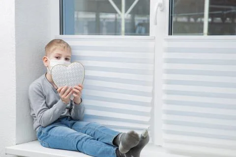 Sad boy sitting on windowsill in protective mask holding heart Stock Photos