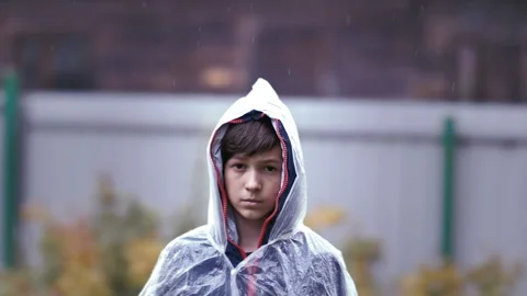 Sad boy standing in the rain with a raincoat, expressing pain on his face Stock Footage 239029929