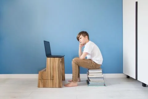 Sad child boy sitting on stack of books with his laptop, studying online at h Stock Photos