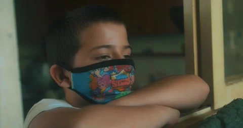Sad child looking outside during the pandemic lockdown with medical mask on Stock Footage 142958530