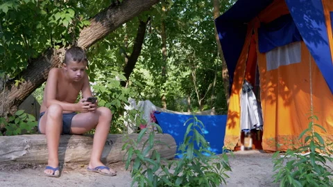 Sad Child Sits on a Log, Playing on a Smartphone in a Forest on Sun near Tent Stock Footage 160851999