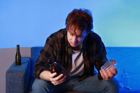 Sad, depressed man looking at an empty bottle of alcohol while sitting on a.. Foto stock