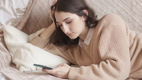 Sad girl chatting using a mobile phone. Young woman lying on the couch at home Stock Footage 165597575