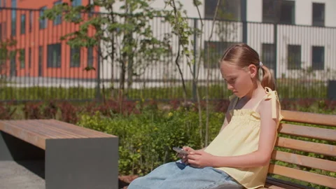 Sad girl using smartphone while sitting on park bench during summer Stock Footage 290825786