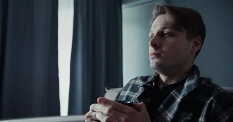 Sad guy with a lifeless look sits on the sofa and drinks a hot drink at home Stock Footage 305787872