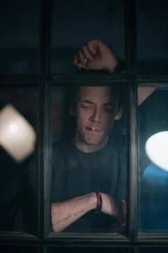 A sad guy smokes outside the window at night. Stock Photos
