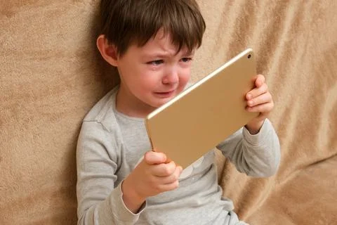 Sad kid crying while holding digital tablet on sofa. Child addiction to gad.. Stock Photos