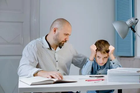 Sad kid sitting at a table next to his father, who is angry at him. Stock Photos
