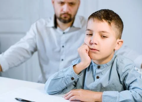 Sad kid sitting at a table next to his father, who is angry at him. Stock Photos