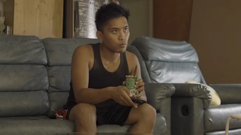 Sad lonely asian man eats beans from can with spoon whilst watching TV Stock Footage 87457359