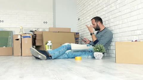 Sad man calculating bills on smartphone sitting on floor at his new home Vídeo Stock 63469834