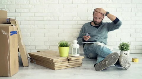 Sad man calculating bills on smartphone sitting on floor at his new home Vídeo Stock 63469890