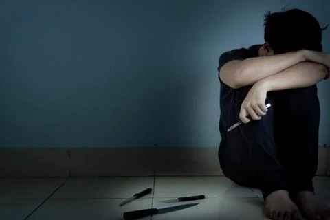 Sad man hug his knee and cry sitting alone and holding a cutter knife with an Stock Photos