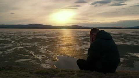 A sad man sits alone on the shore of a lake at sunset. Video stock 333205974