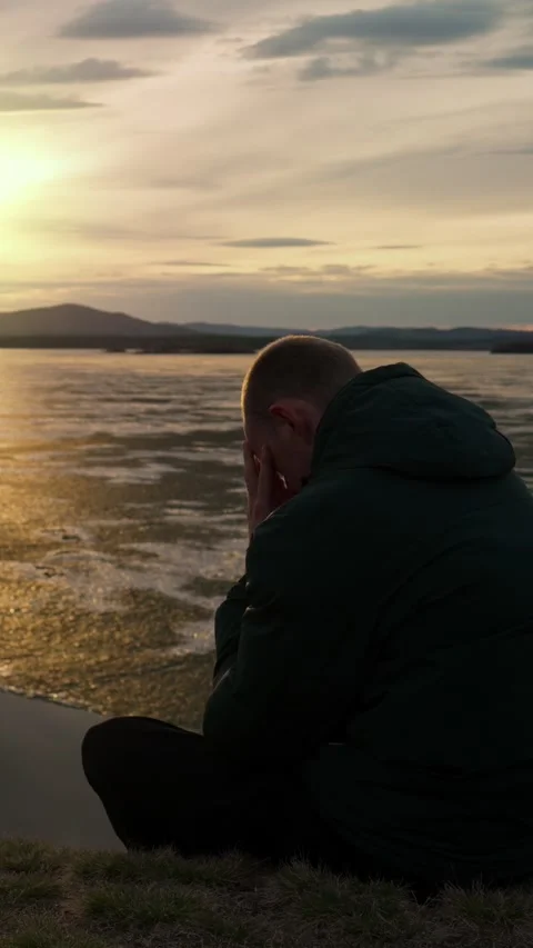 A sad man sits alone on the shore of a lake at sunset. Stock-Footage 344021336