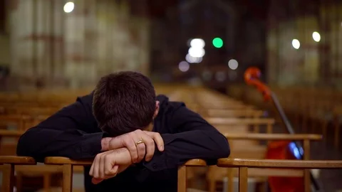 Sad man is sitting bowing head on crossed hands on chair in empty hall of church Stock Footage 99576011