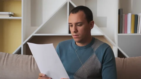 Sad man sitting on couch at home reads received bad news holds documents paper Stock Footage 134873464