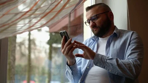 Sad man by the window and texting on smartphone 스톡 동영상 71657113