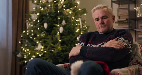 A sad middle-aged man with gray hair is sitting on chair near Christmas tree Stock Footage 210573131