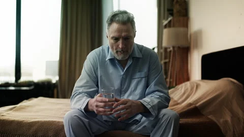 Sad middle-aged man sitting on bed and taking pill with water, health problems Stock Footage 321615564