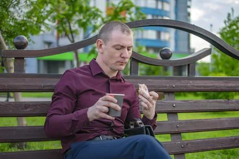 Sad office worker eats a sweet donut with coffee in a city park Stock Photos