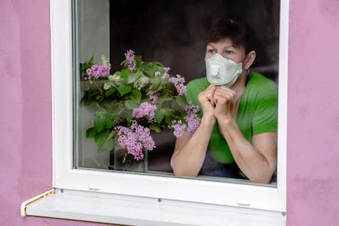 A sad older woman in a protective mask looks out the window, waiting for the  Stock Photos