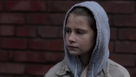 Sad pensive homeless refugee hooded child girl dark portrait against brick wall Stock Footage