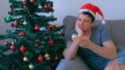 Sad single man in red cap boring sitting on couch near Christmas tree at home. Stock Footage 137461522