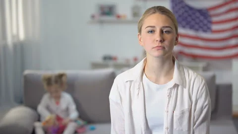 Sad single mother looking at camera daughter playing on sofa against flag of USA Stock Footage 108452728