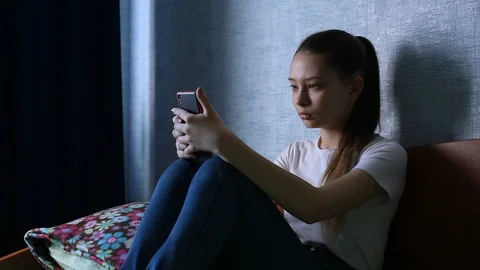 A sad teenage Girl sits on the bed, puts down her phone and Looks out the window Stock Footage