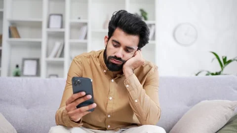 Sad young handsome bearded man bored browsing using smartphone while sitting  Stock Footage 252216110