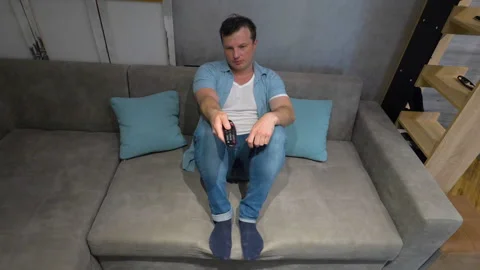 Sad young man watching TV, guy changing TV channels with remote control Stock Footage 323921149