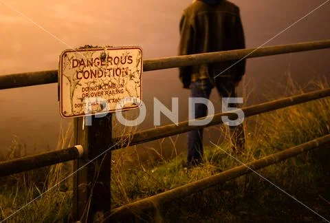 Sad/depressed man standing on edge of cliff behind a danger sign Stock Photos