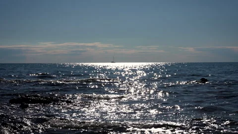 Sailboat Passing Under Bright Sun Over Gentle Waves and Rocky Shore Stock Footage 341630285