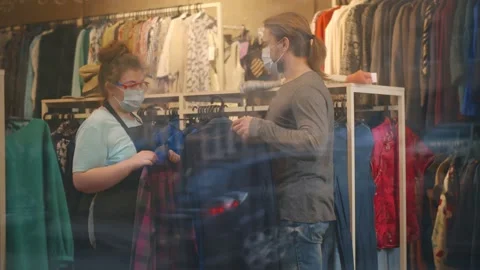 Sales assistant in protective mask helping male customer to buy clothes in Stock Footage 143568744