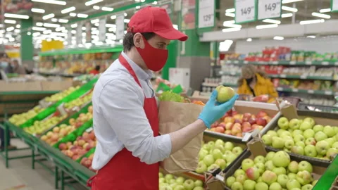 Salesman in red face mask standing in grocery store selecting fresh apples Stock Footage 149106495