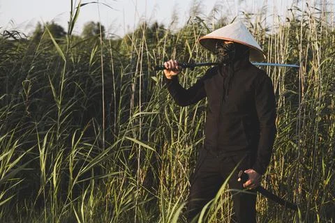 A samurai wearing a mask, conical hat and black clothes holds a katana over his Stock Photos