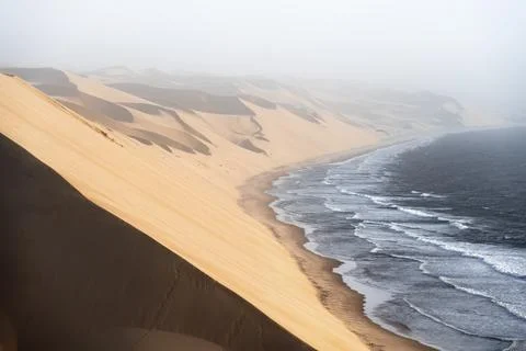 Sand dunes with wave patterns and sea in evening light, wind blowing sand across Stock Photos